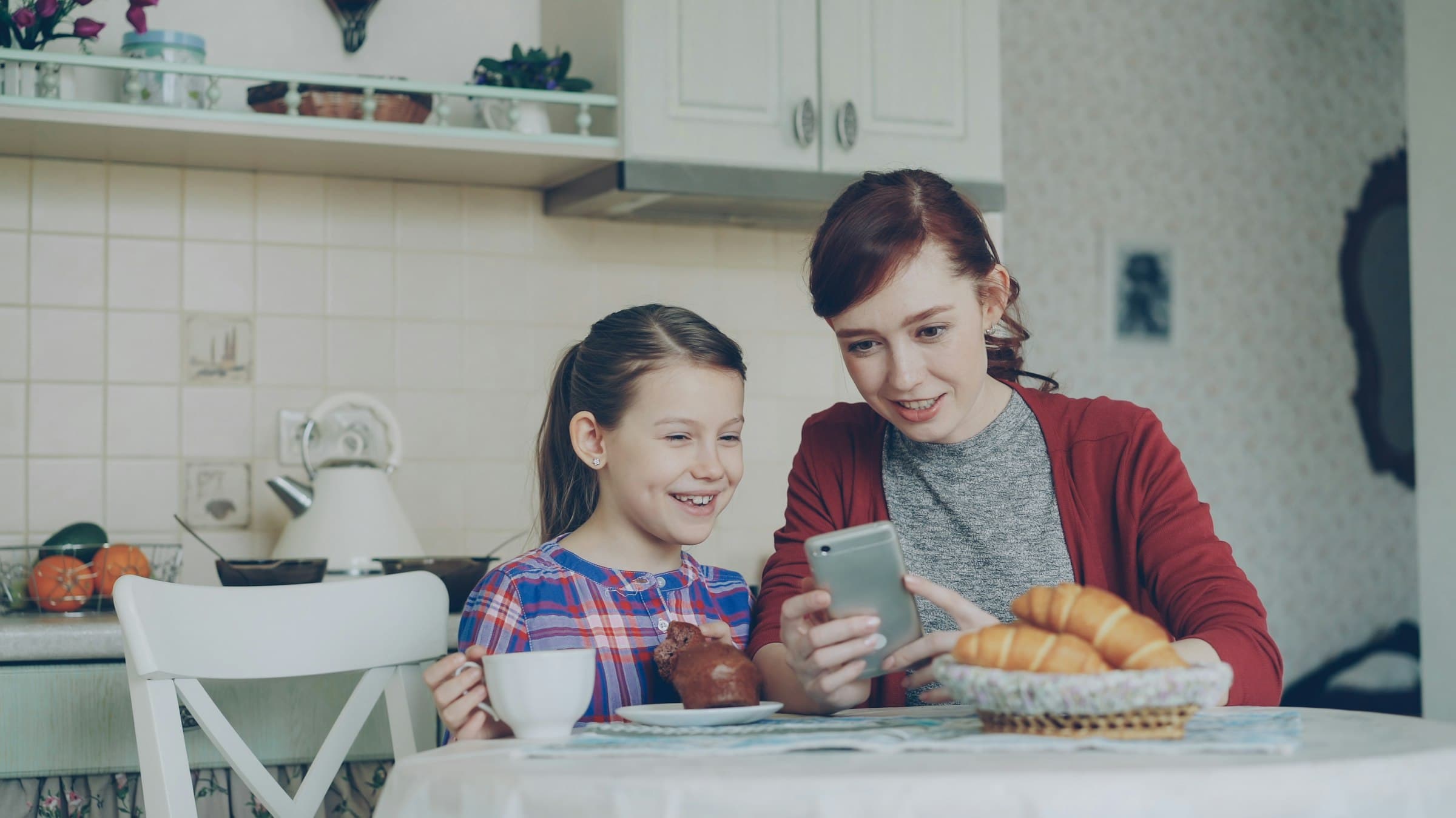 A mother and daughter laughing together over a phone at the kitchen table